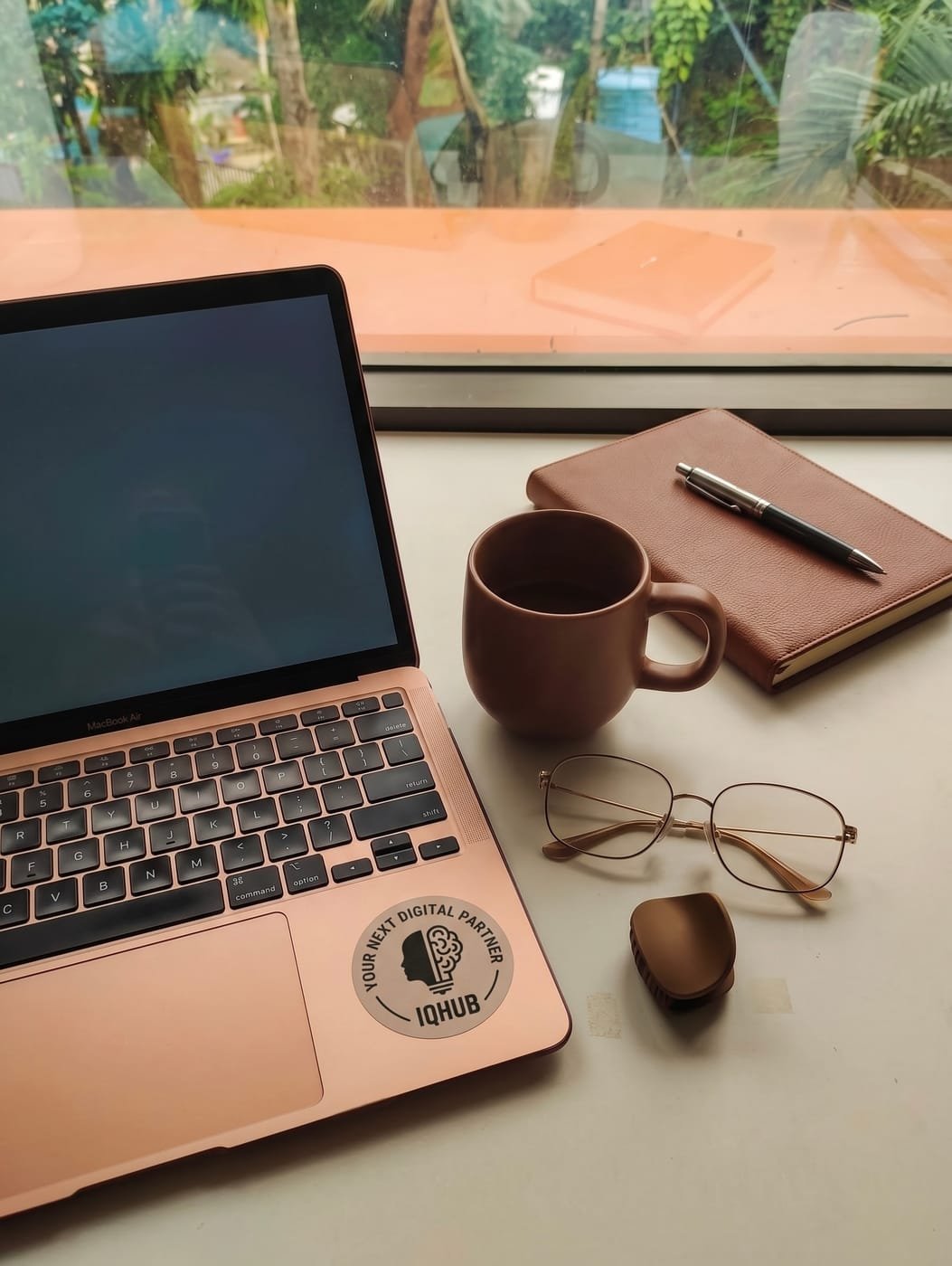 A laptop with an IQhub sticker, notebook and coffee on a sunlit desk